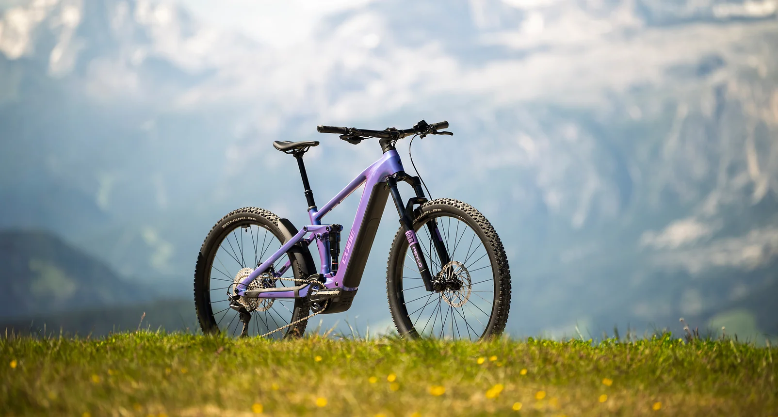 Cube Stereo Hybrid ONE44 Pro 800 in blueiris´n´purple, das ganze Rad in der Seitenansicht auf einer Bergwiese mit gelben Blüten, im Hintergrund unscharf ein Alpenpanorama mit Schneeresten.