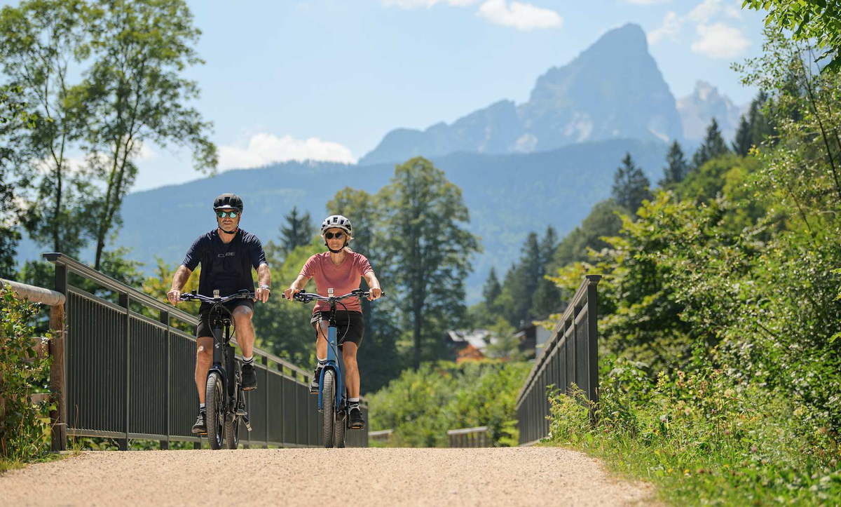 CUBE Kompakt E-Bikes 2027: zwei Fahrer auf einer Brücke vor Bergpanorama