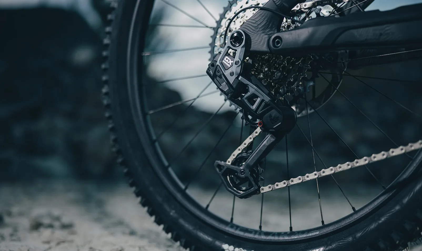 Sram GX Eagle AXS Schaltwerk am CUBE Stereo Hybrid ONE77 HPC TM 2027 im Gelände, am Käfig die Aufschrift GX, dahinter Kassette, Speichen und Reifen, im Hintergrund unscharfe Felsen