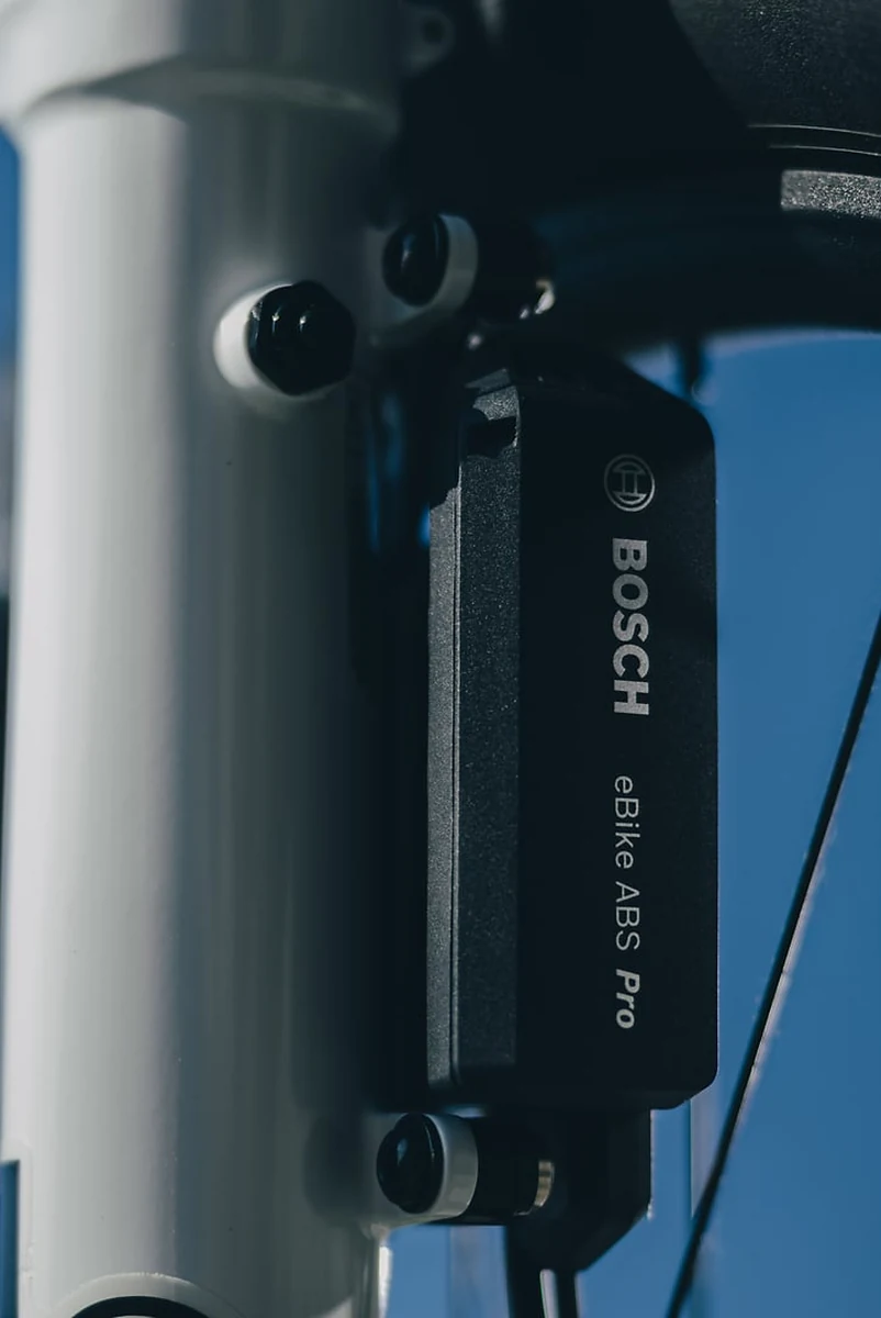 Bosch eBike ABS Pro am CUBE Stereo Hybrid ONE77 AT 2027 in carbon´n´white, schwarzes Modul mit dem Schriftzug BOSCH und eBike ABS Pro, außen am weißen Gabelholm verschraubt, dahinter blauer Himmel