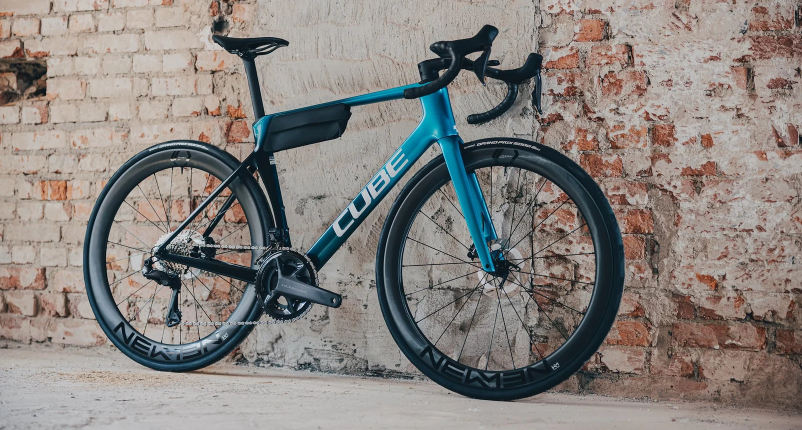 CUBE Agree C:62 Race 2027 in chromeblue´n´liquidblue im Seitenprofil vor einer Ziegelwand, tiefe Newmen-Laufräder und eine schwarze Rahmentasche im Rahmendreieck