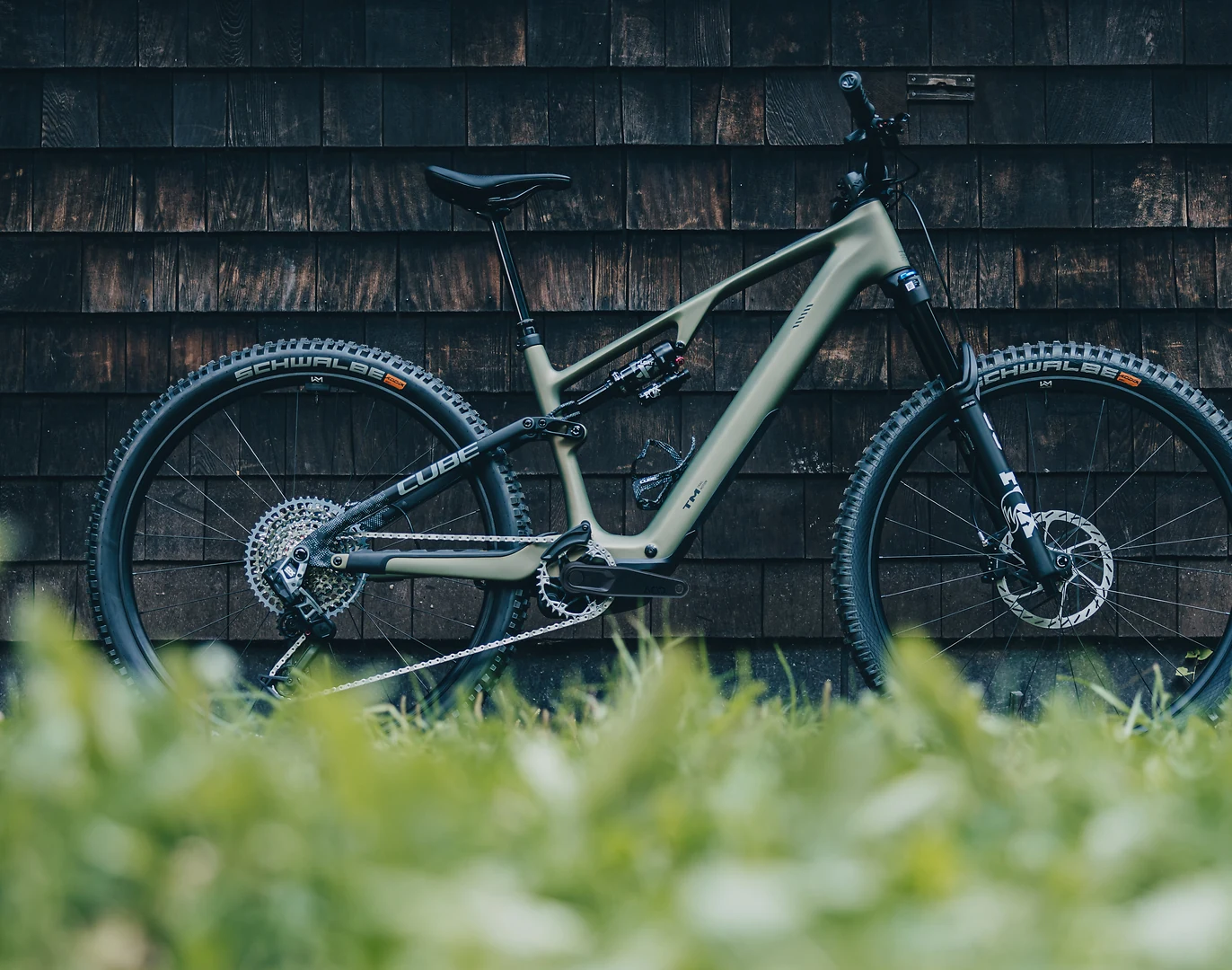 CUBE AMS Hybrid ONE44 C:68X TM 2026 in reedgreen´n´matrix, das ganze Rad in der Seitenansicht vor einer dunklen Holzschindelwand, im Vordergrund unscharfes grünes Gras, auf der Sitzstrebe der Schriftzug CUBE, Schwalbe-Reifen