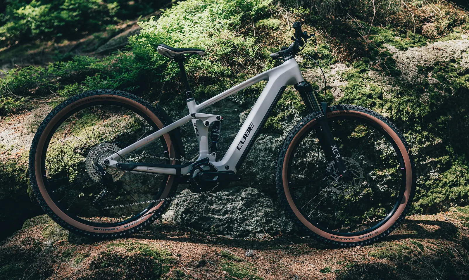 CUBE Stereo Hybrid ONE22 Pro 800 2027 in foggrey´n´sparklerose in der Seitenansicht an einem bemoosten Felsen im Wald, Schwalbe-Smart-Sam-Reifen mit brauner Flanke, Bosch-Motor am Tretlager
