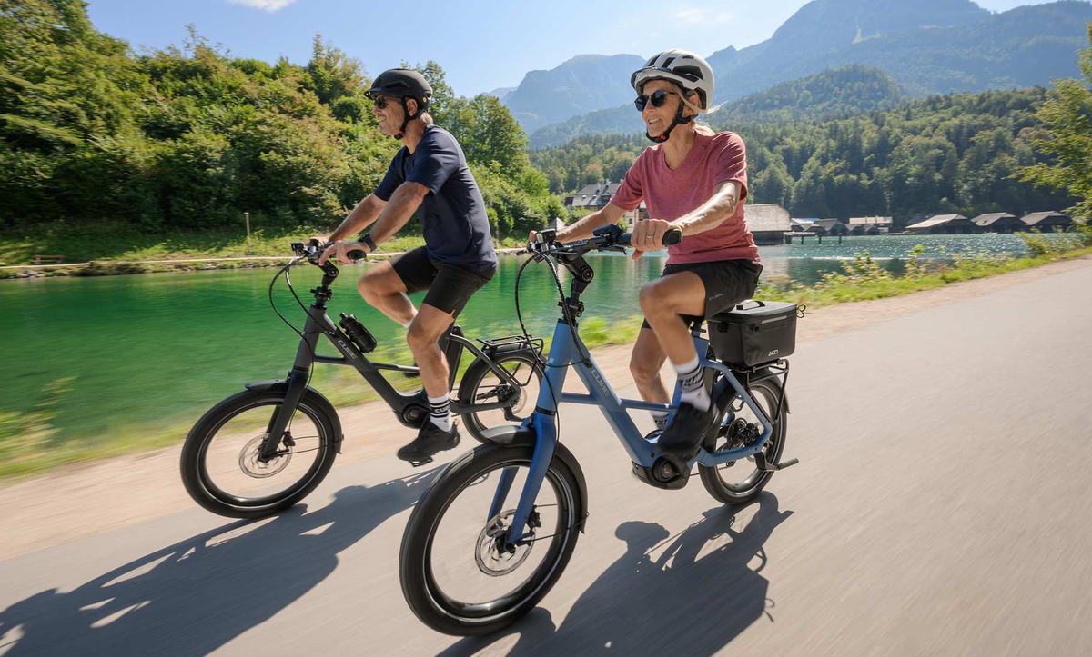 CUBE Compact Hybrid 2027: zwei Fahrer nebeneinander auf einem Uferweg vor Bergen und türkisem See