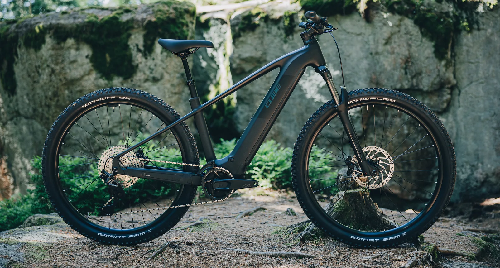 CUBE Reaction Hybrid Pro 800 2027 in silverdust´n´prism im Seitenprofil vor einer bemoosten Felswand, dunkelgrauer Rahmen, starrer Hinterbau ohne Dämpfer, Federgabel vorn, Schwalbe Smart Sam Reifen, Boden aus Erde und Wurzeln.