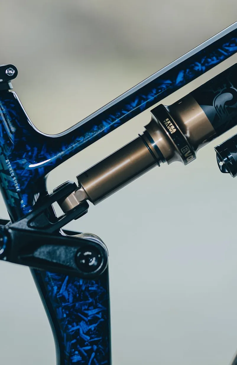 Dämpfer des Cube Stereo Hybrid ONE44 HPC SLT 800 in splinternavy´n´prizm aus der Nähe. Der goldfarbene Dämpferkörper sitzt schräg im dunkelblau marmorierten Carbon-Hinterbau, links davon die Umlenkung mit Lagerschrauben.