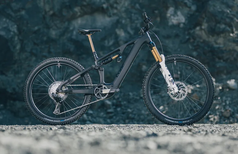 CUBE AMS Hybrid 177 C:62 AT 600X 2027 in carbon´n´white im Seitenprofil vor einer dunklen Felswand, schwarzer Rahmen, Federgabel mit weißen Tauchrohren und goldfarbenen Standrohren, Dämpfer im Rahmendreieck, Schotterboden.