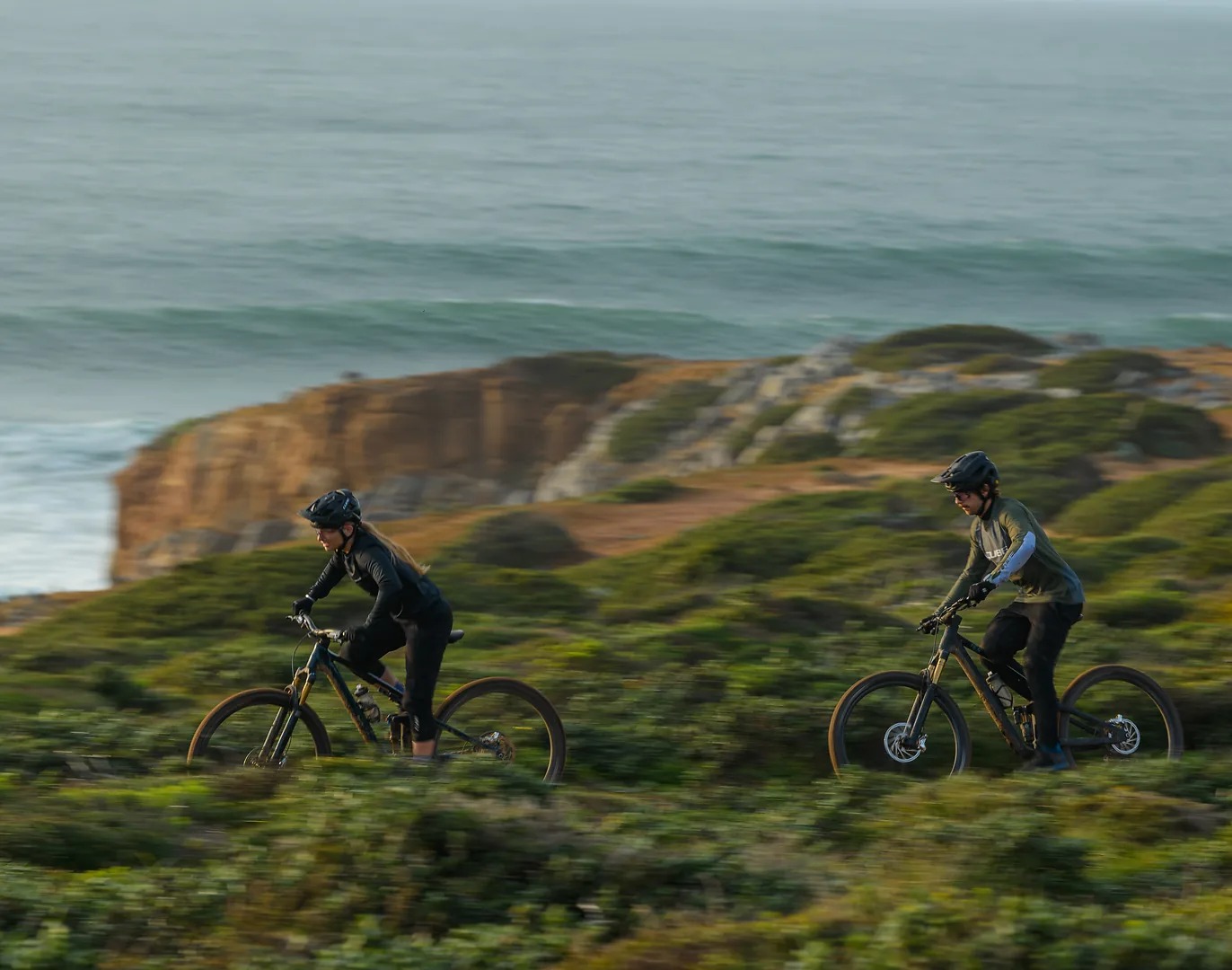 Zwei Fahrende auf vollgefederten CUBE E-Mountainbikes auf einem Küstentrail, links eine Frau in schwarzer Kleidung, rechts ein Mann im olivgrünen Trikot, dahinter Buschland, eine Felsküste und das Meer.