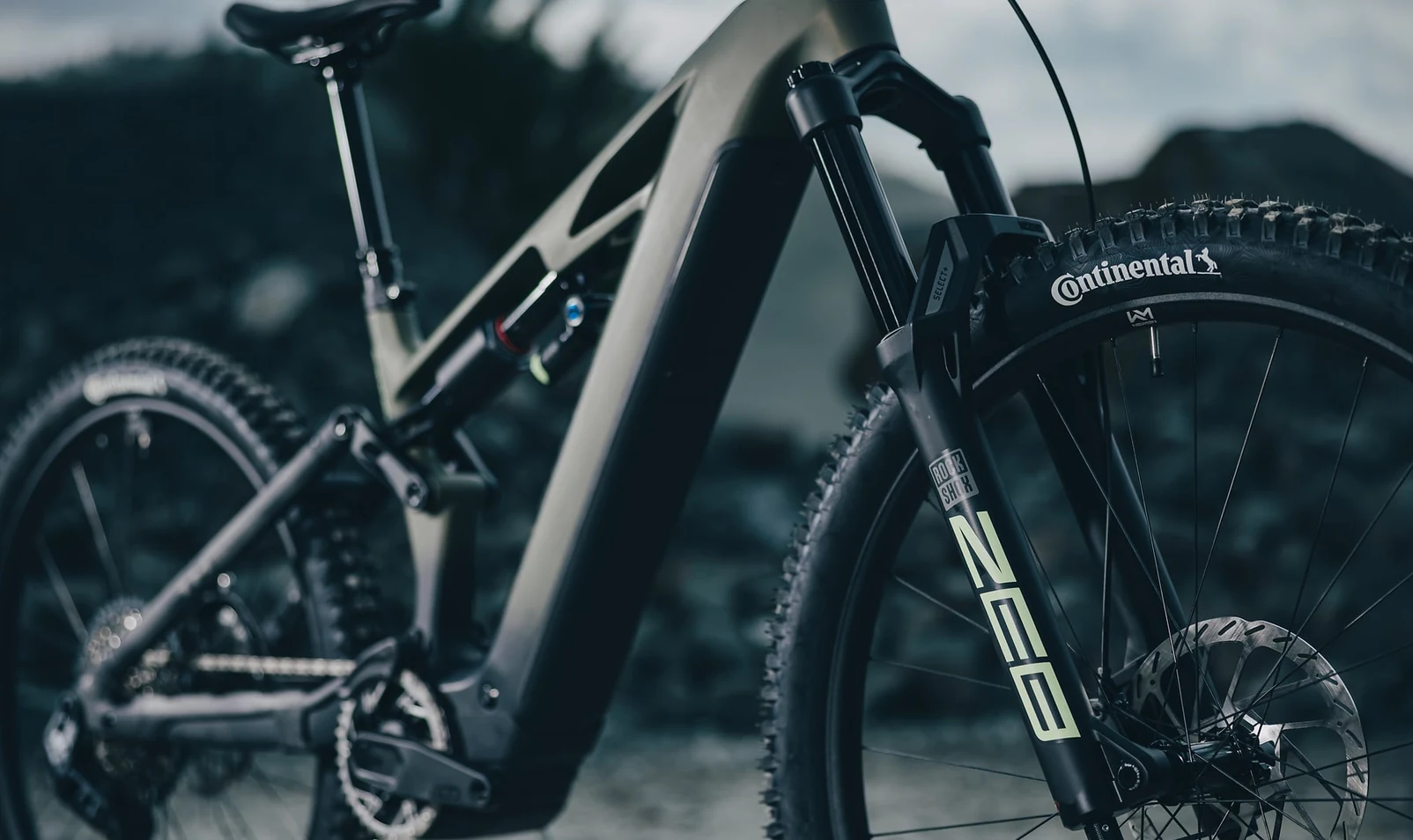 Vorderpartie eines CUBE Stereo Hybrid ONE77 TM 2027 in reedgreen´n´gills, RockShox-ZEB-Federgabel mit grünem Schriftzug und der Kennzeichnung SELECT+ am Gabelkopf, davor ein Continental-Reifen, im Hintergrund unscharf die Geröllhalde eines Steinbruchs