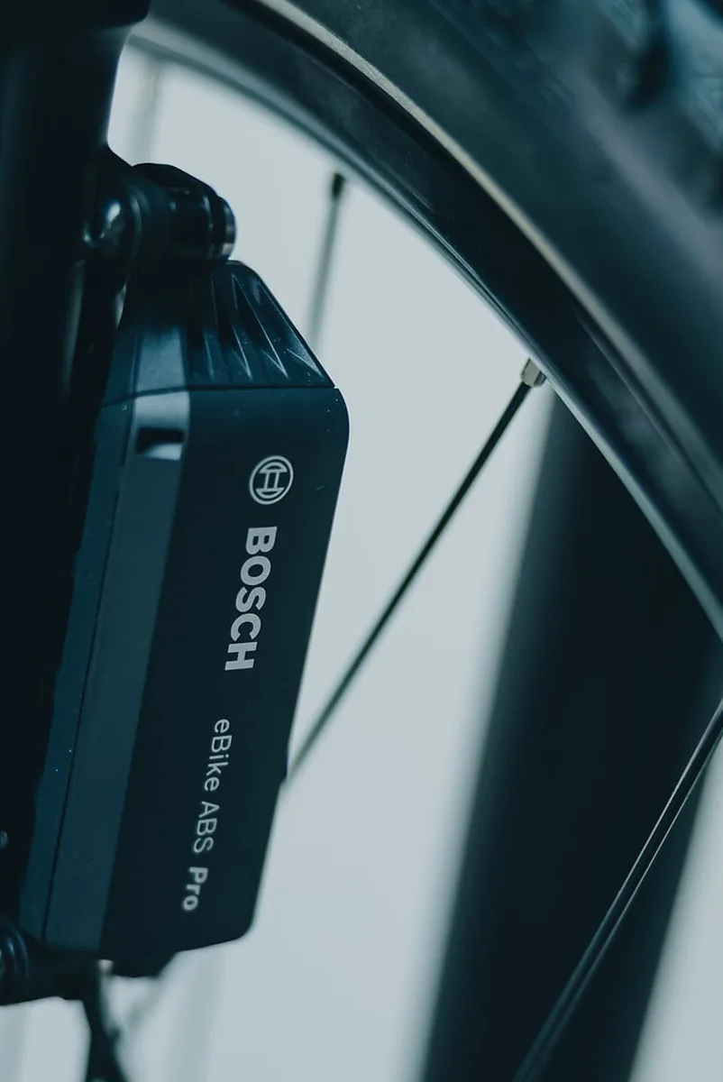 Schwarzes Bosch-eBike-ABS-Pro-Modul, seitlich am Gabelholm des Cube Stereo Hybrid ONE44 SLX 800 in smaragdgrey´n´chrome verschraubt. Im Hintergrund unscharf die Speichen und der Stollenreifen des Vorderrads.