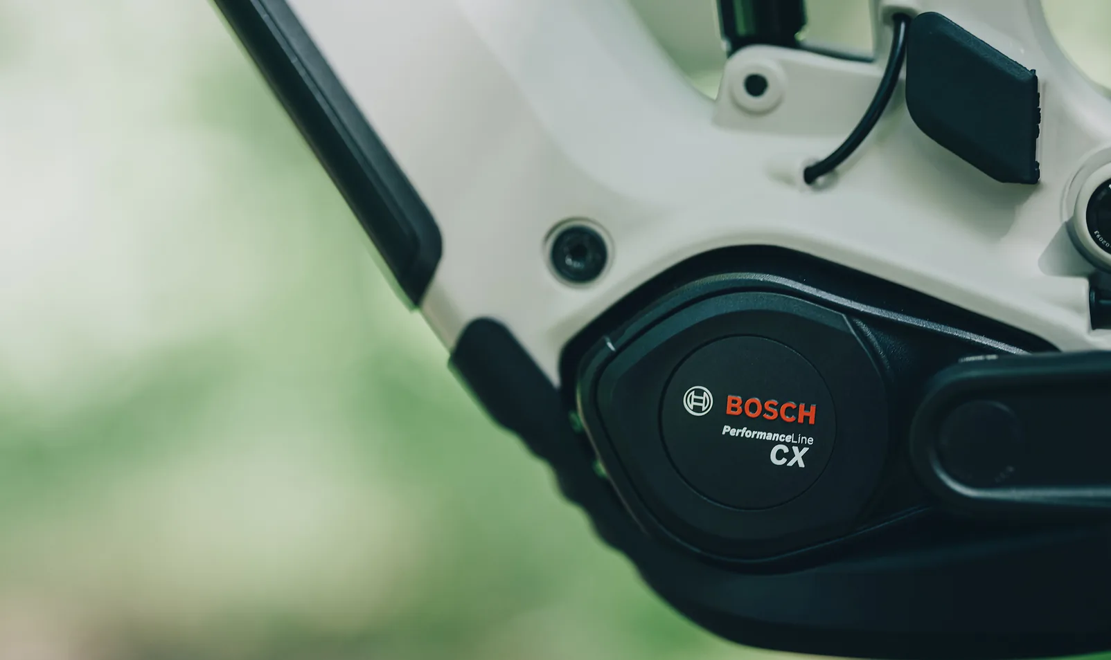 Motordeckel am CUBE Stereo Hybrid ONE44 SLX 800 2027 in oatgrey´n´grey aus der Nähe, auf dem schwarzen runden Deckel steht Bosch Performance Line CX, darüber der helle Rahmen mit der Akkuabdeckung, im Hintergrund unscharfes Grün.