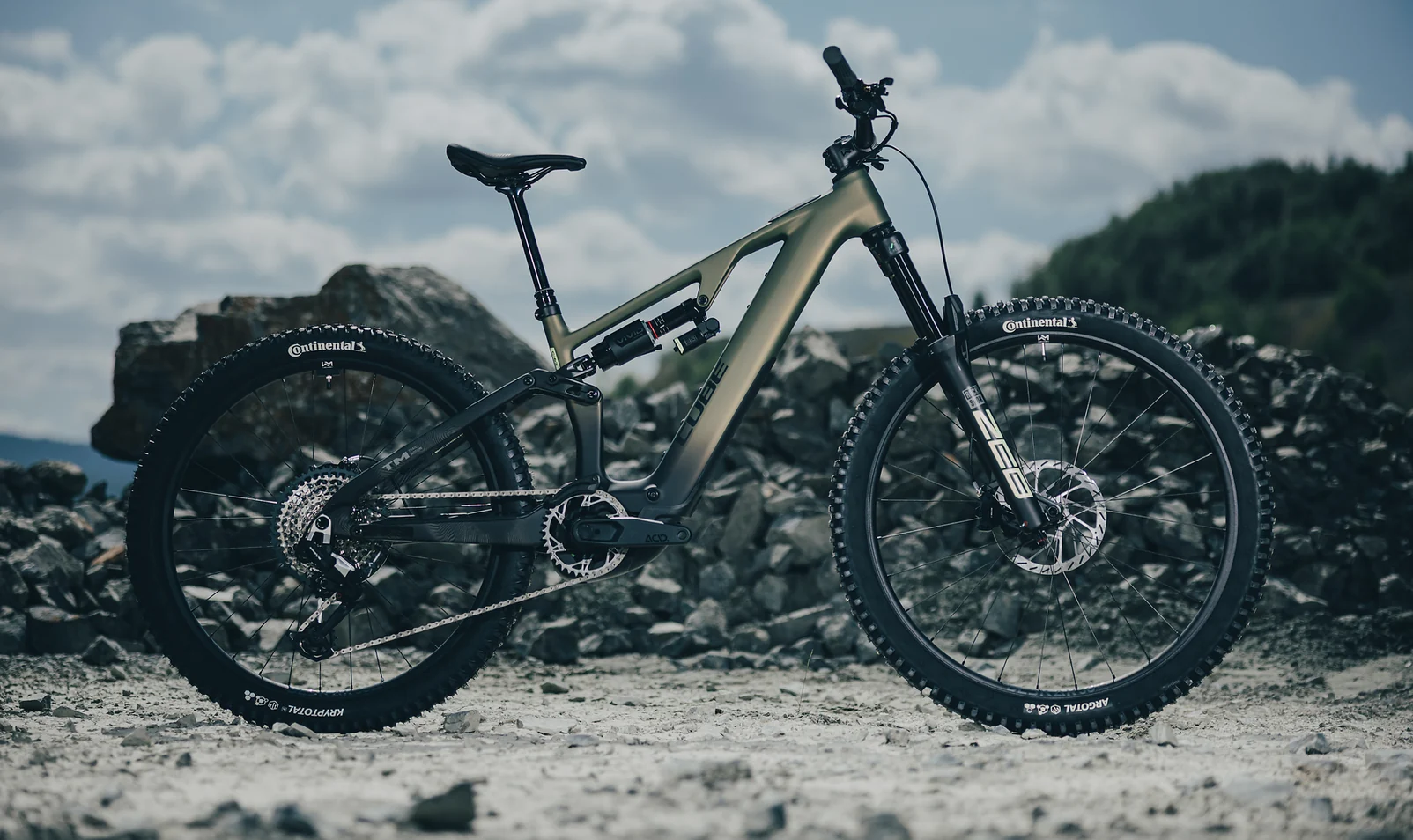CUBE AMS Hybrid 177 C:62 TM 2027 in reedgreen´n´gills, das ganze Rad in der Seitenansicht auf hellem Geröll, auf dem Unterrohr der Schriftzug CUBE, an der schwarzen Federgabel der Schriftzug ZEB, Continental-Reifen, dahinter Felsblöcke, Nadelwald und bewölkter Himmel