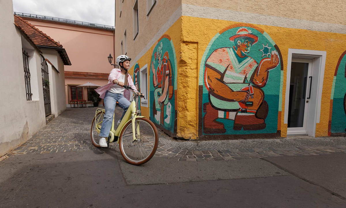 CUBE Aruba Hybrid 2027: Fahrt durch eine gepflasterte Altstadtgasse an einem bunten Wandbild vorbei