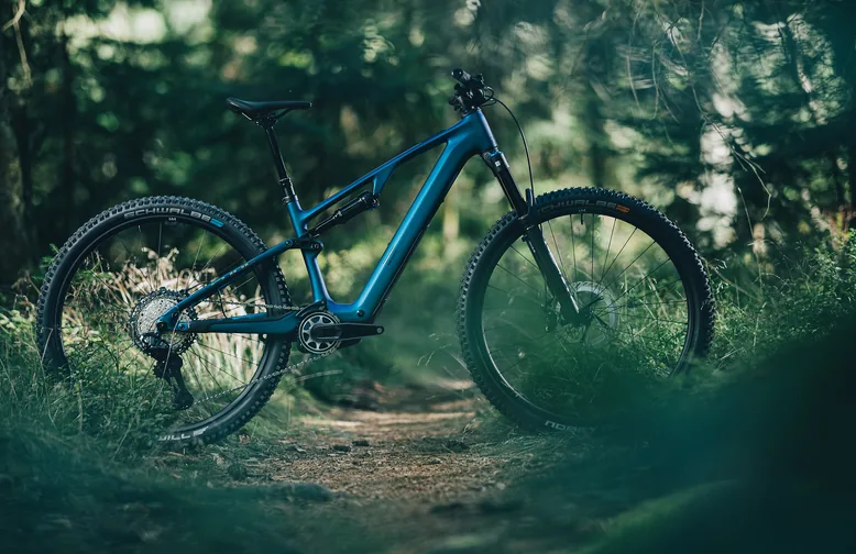CUBE AMS Hybrid ONE44 C:68X SLX 2026 in nebula´n´black, das ganze Rad in der Seitenansicht nach rechts auf einem schmalen Waldweg, ringsum Gras und Nadelbäume, Schwalbe-Reifen