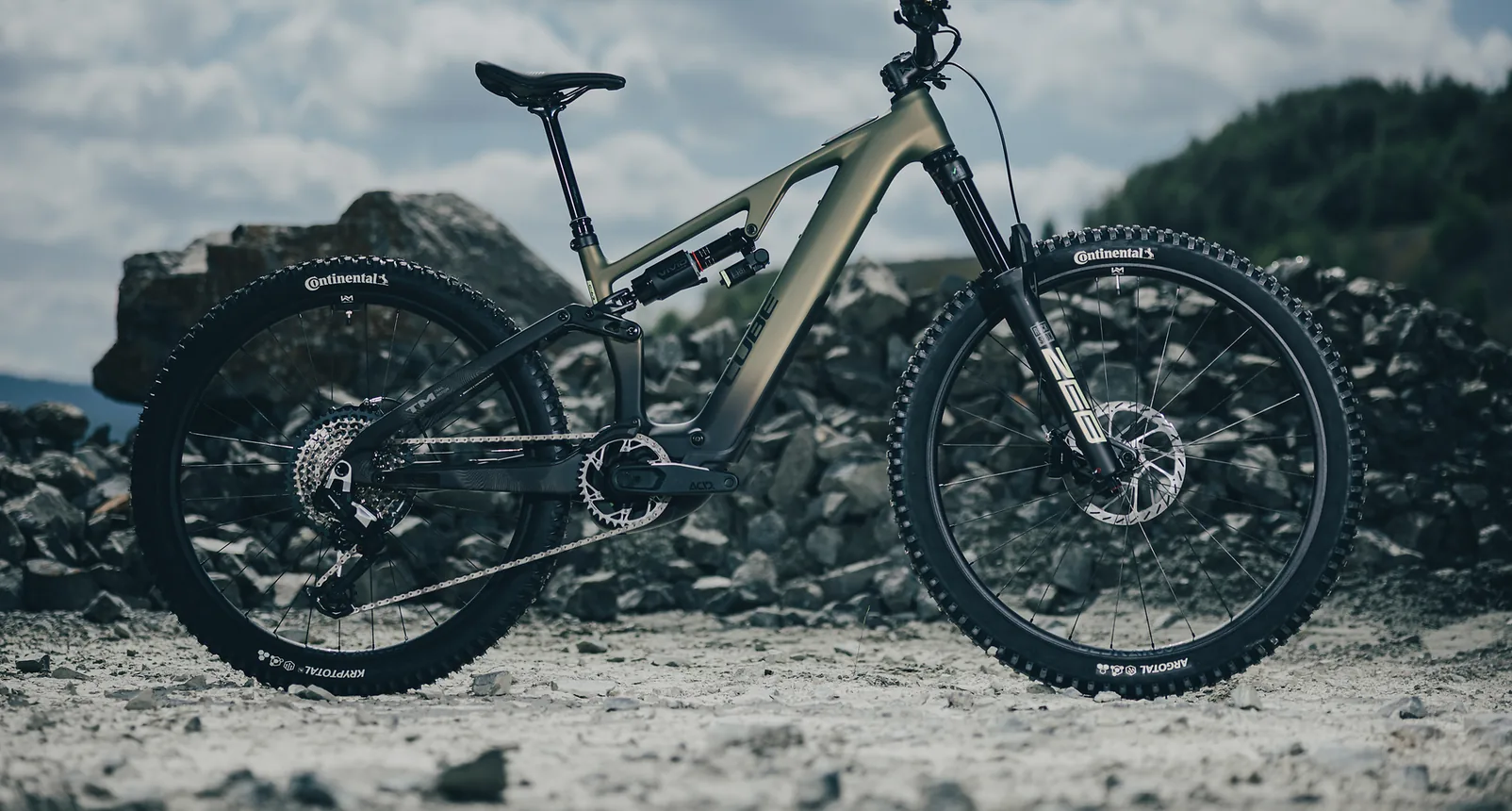 CUBE AMS Hybrid 177 C:62 TM 600X 2027 in reedgreen´n´gills im Seitenprofil vor einer Halde aus grauem Gestein, olivgrüner Rahmen mit schwarzem Unterrohr, RockShox Vivid Dämpfer und ZEB Federgabel, Continental Reifen, dahinter Wolkenhimmel und ein bewaldeter Hang.