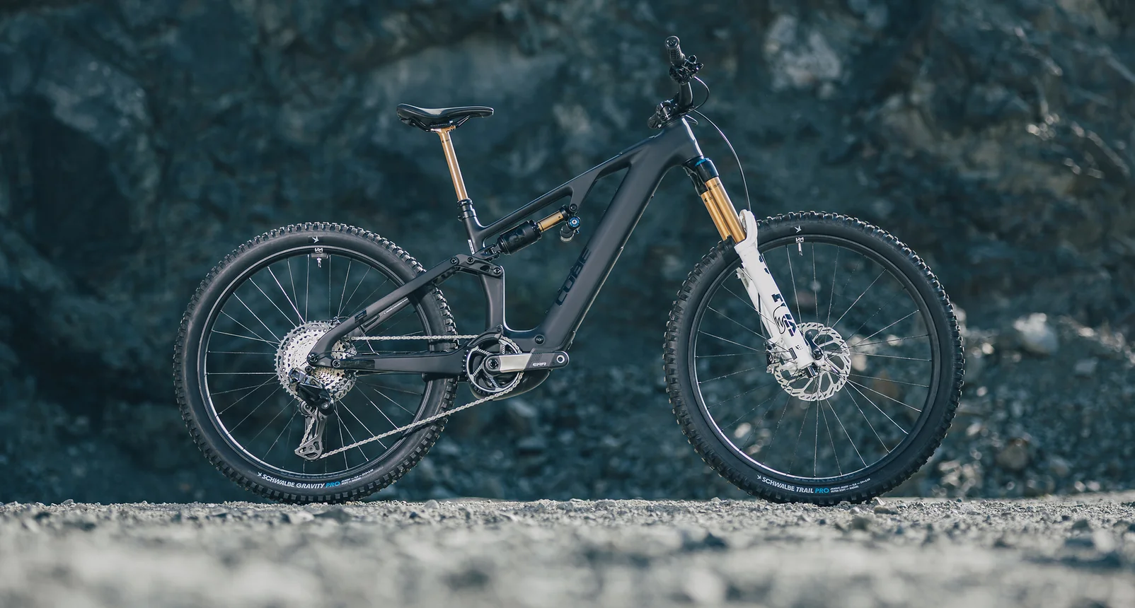 Cube AMS Hybrid 177 AT 600X in carbon´n´white, das ganze Rad in der Seitenansicht auf grobem Schotter vor einer dunklen, feuchten Felswand in einem Steinbruch, weiss lackierte Fox-Gabel mit Schwalbe-Bereifung.