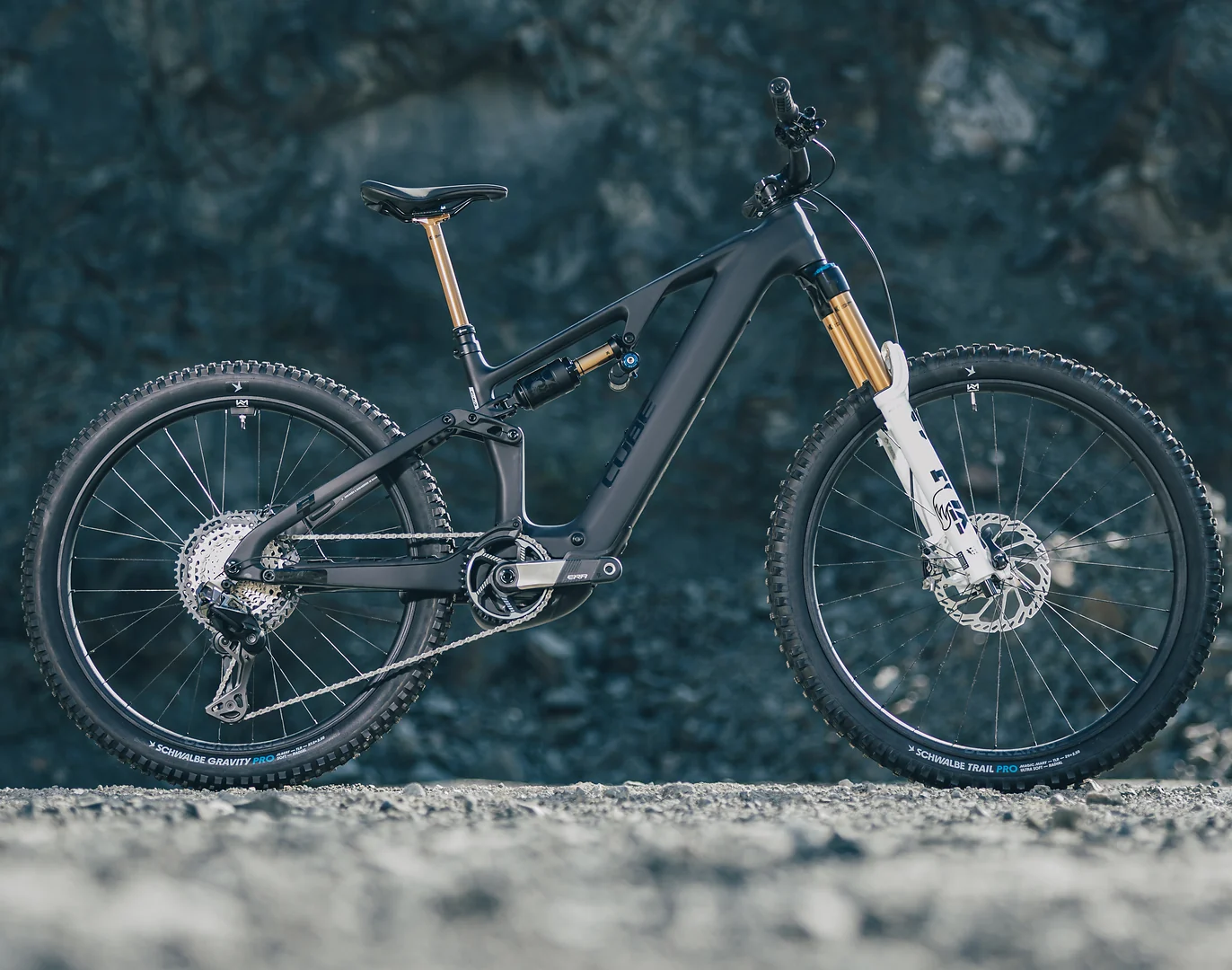 CUBE AMS Hybrid 177 C:62 AT 2027 in carbon´n´white, das ganze Rad in der Seitenansicht auf Schotter vor einer dunklen Felswand, weiße Federgabel mit goldenen Standrohren, auf dem Unterrohr der Schriftzug CUBE, Schwalbe-Reifen