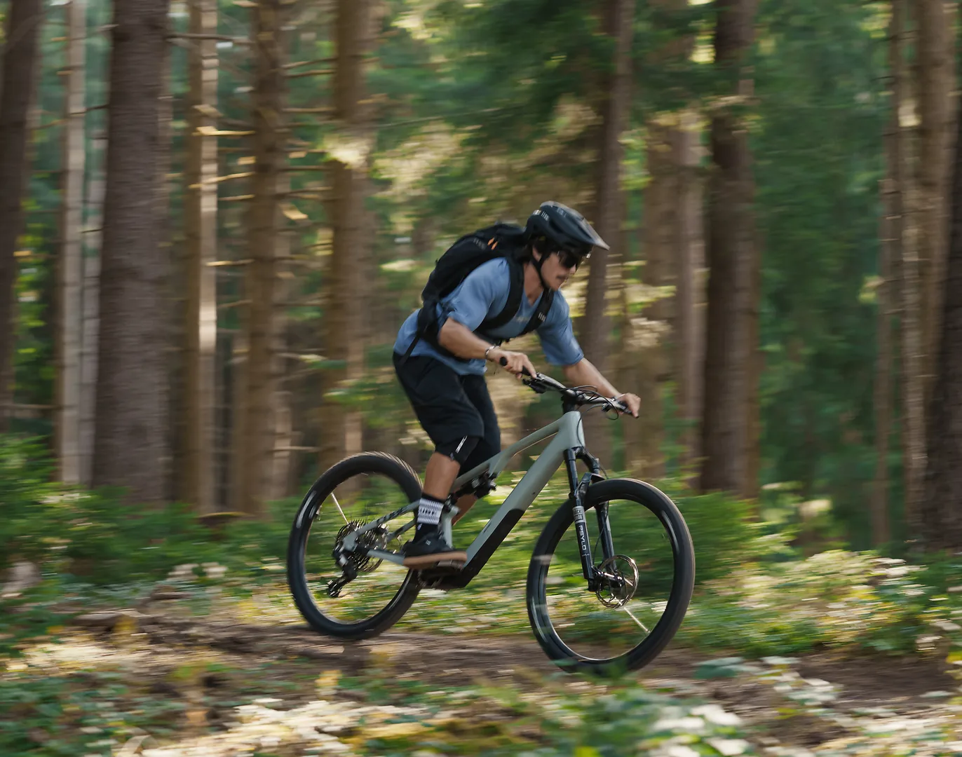 Fahrer auf einem vollgefederten E-Mountainbike in schneller Fahrt auf einem Waldweg zwischen Nadelbäumen, hellgrauer Rahmen, Halbschalenhelm und Rucksack, im Hintergrund Bewegungsunschärfe